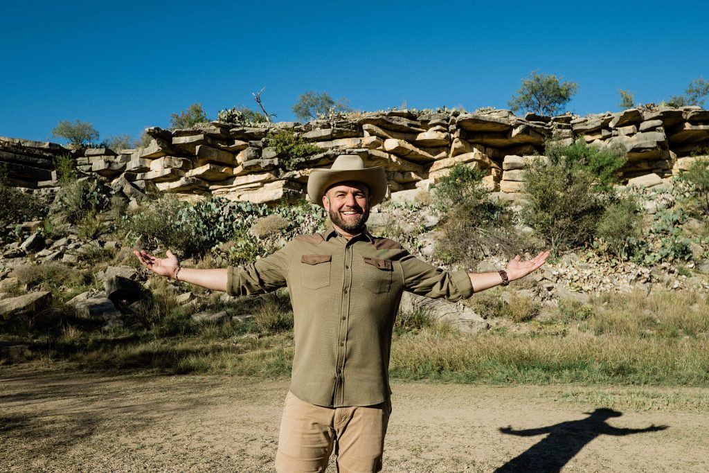 A man in a cowboy hat and forest green shirt stands with his arms outstretched in front of a rock formation