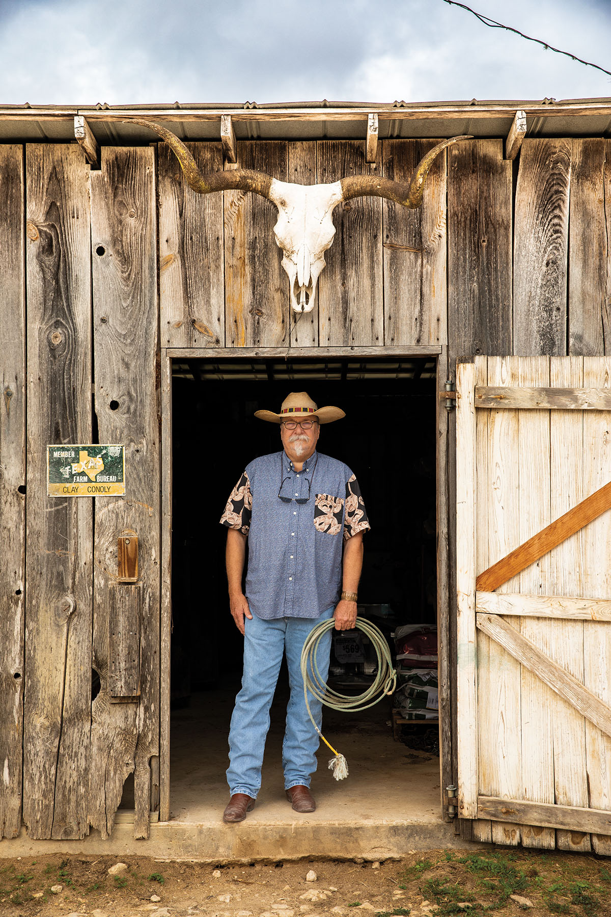 A man in a Western shirt and hat stands in the doorway of a wooden structure holding a rope