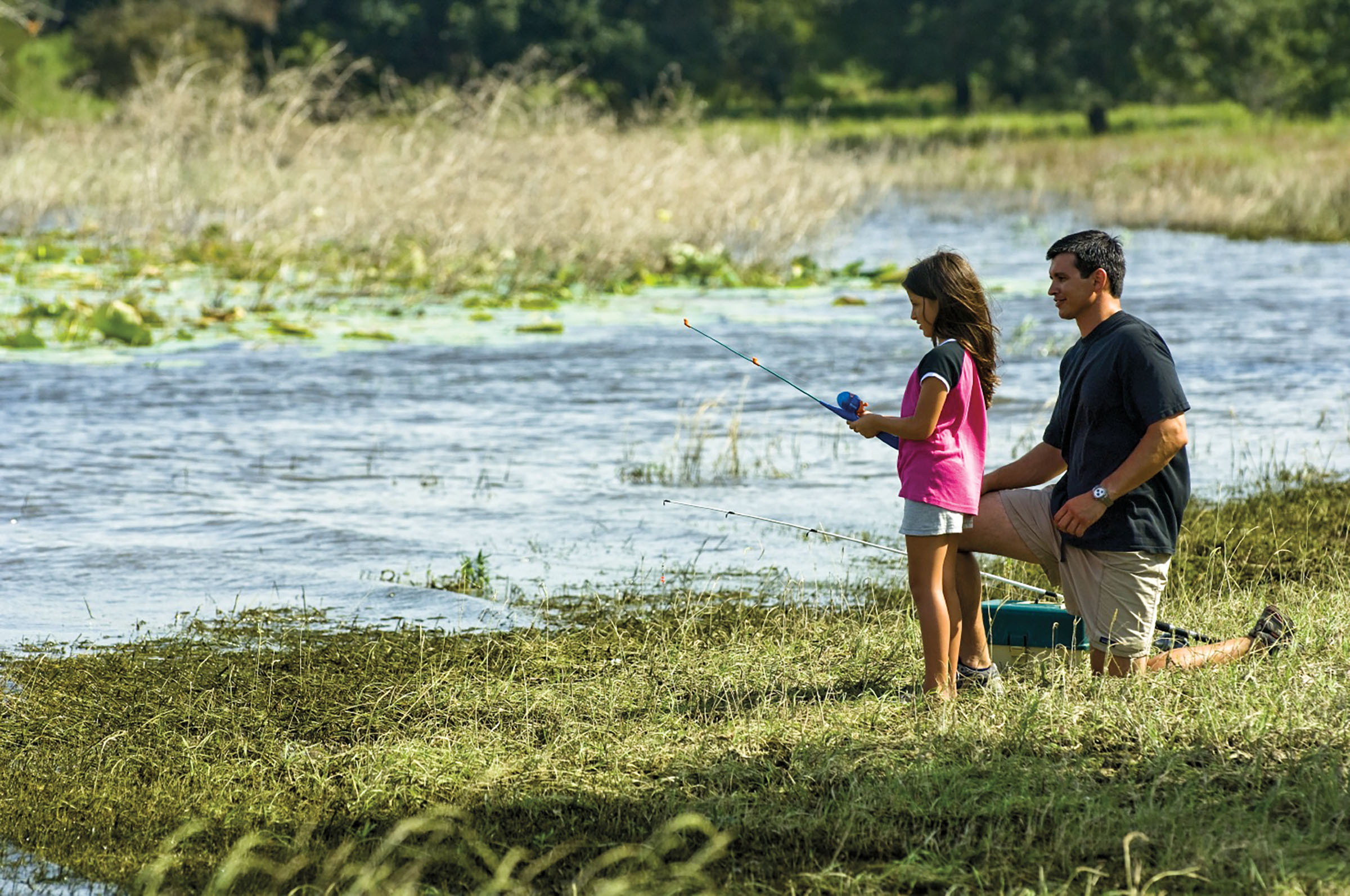 A man and a young woman kneel with fishing rods next to a small body of water and lots of green grass