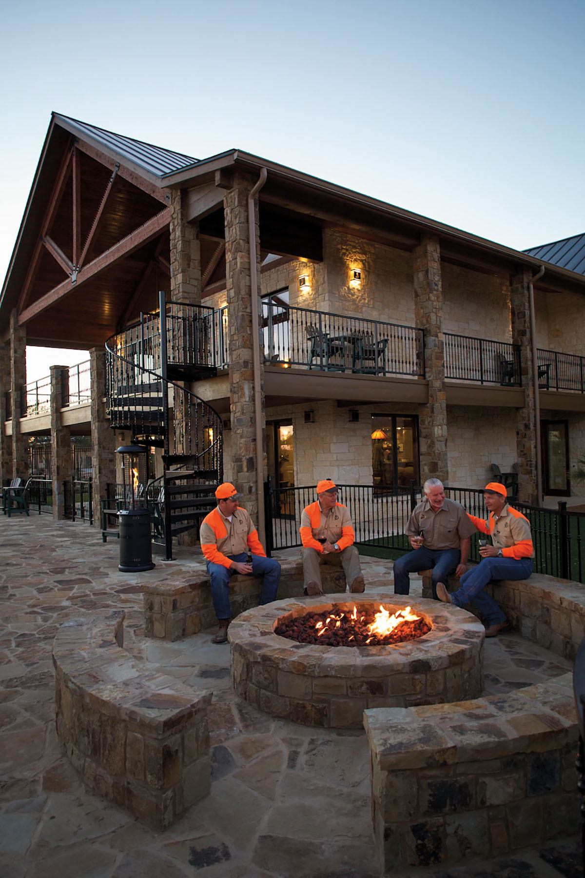 A group of people in orange reflective vests sit around a firepit outside a large lodge