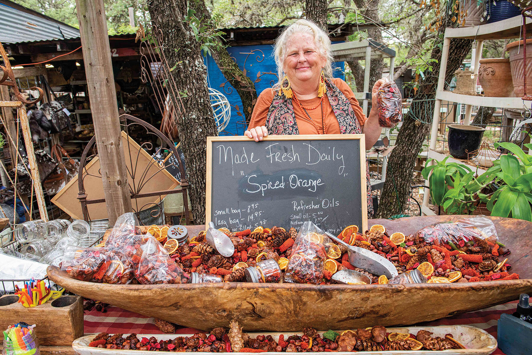 A woman stands behind a small chalkboard reading Made Fresh Daily: Spiced Orange and an assortment of potpurri