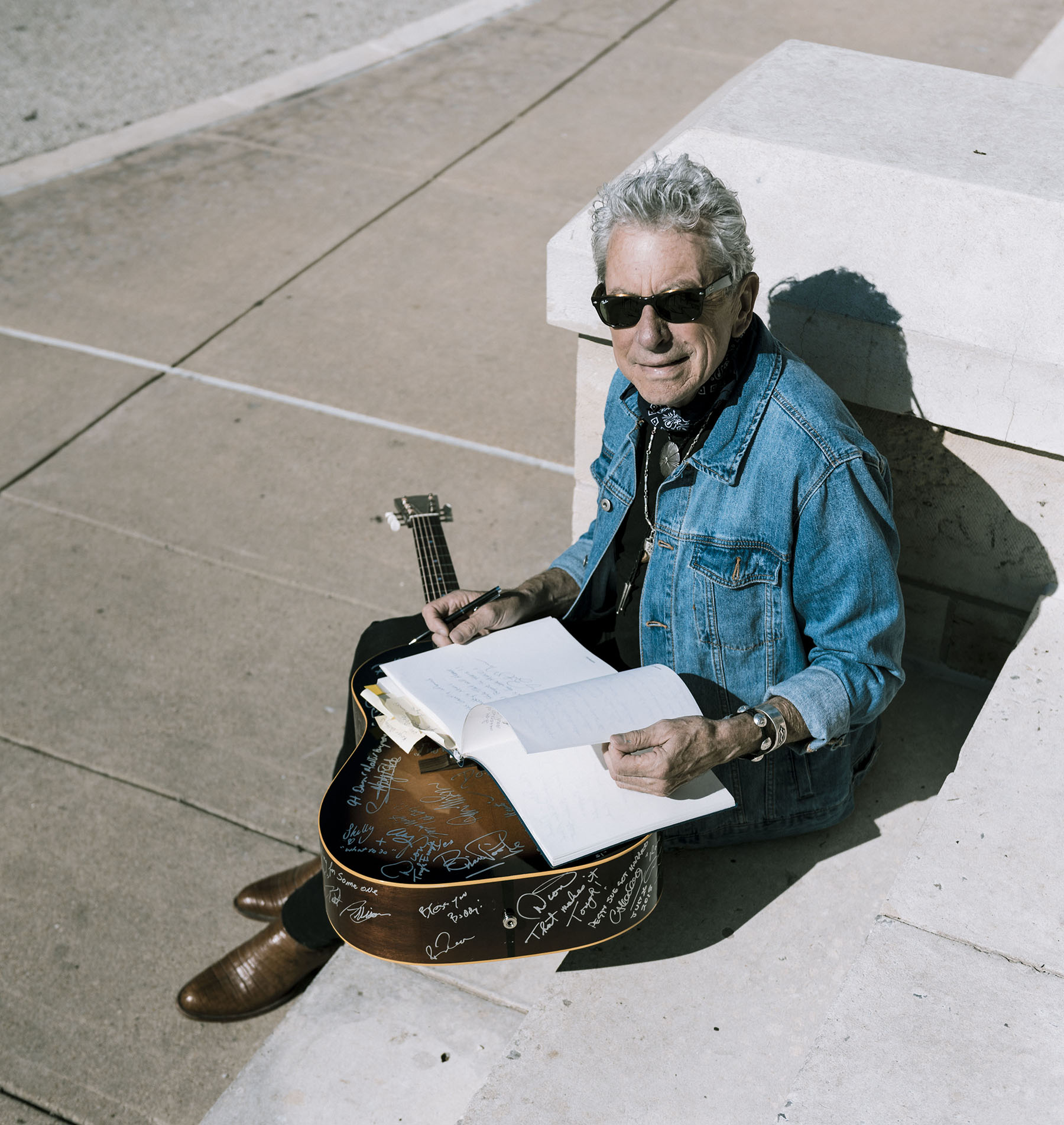 A man in a denim jacket and sunglasses sits on conrete steps with a guitar on his lap and book open over the guitar