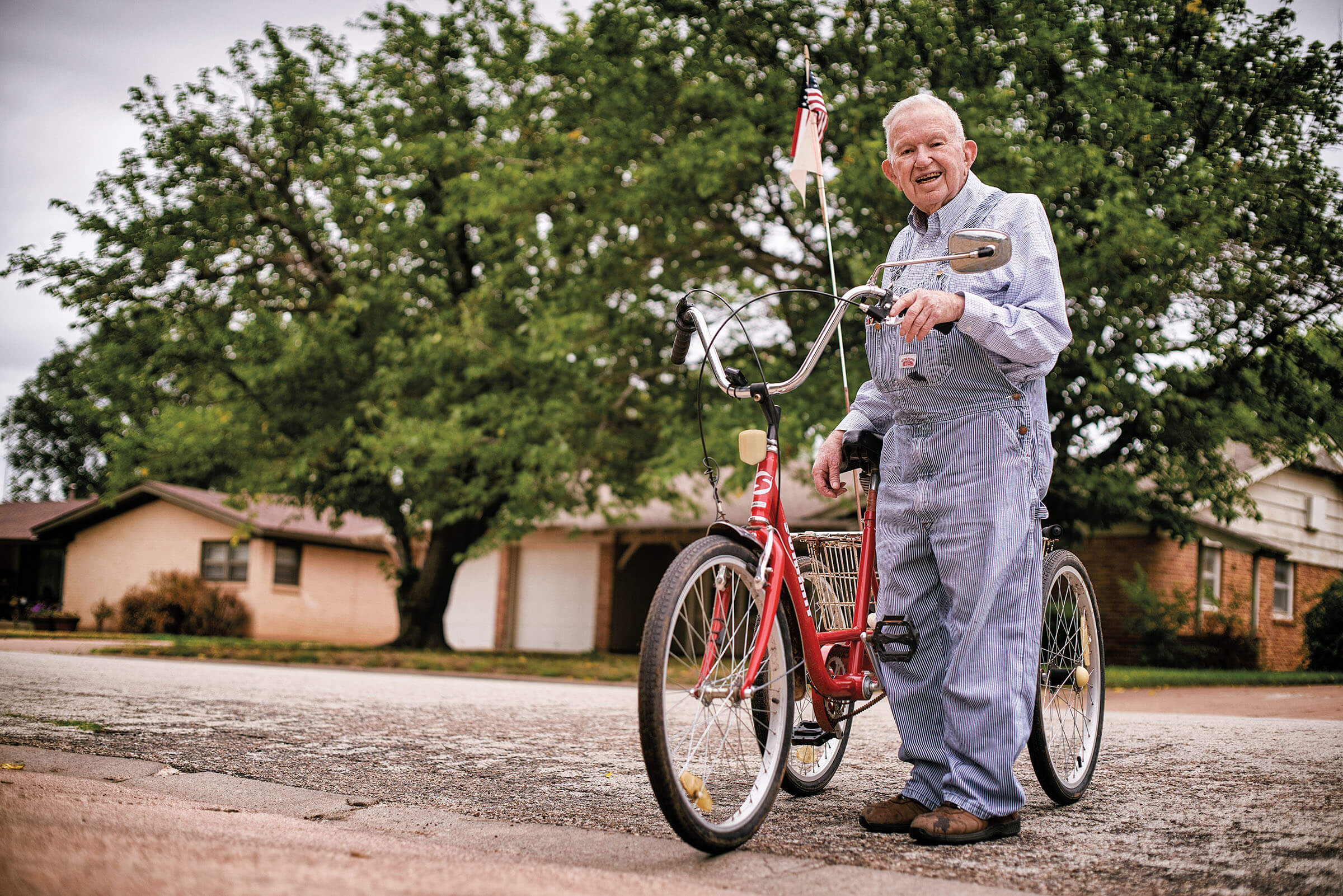 A man in pinstripe overalls stands next to a bright red three-wheeled bicycle with American and Texas flags on the back