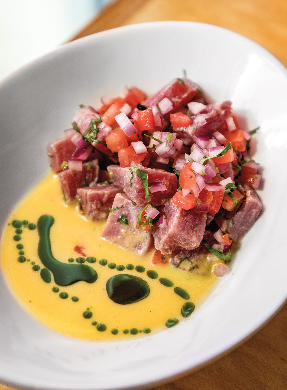 A white plate with a large portion of pink, red and white ceviche next to a bright yellow sauce with green accents