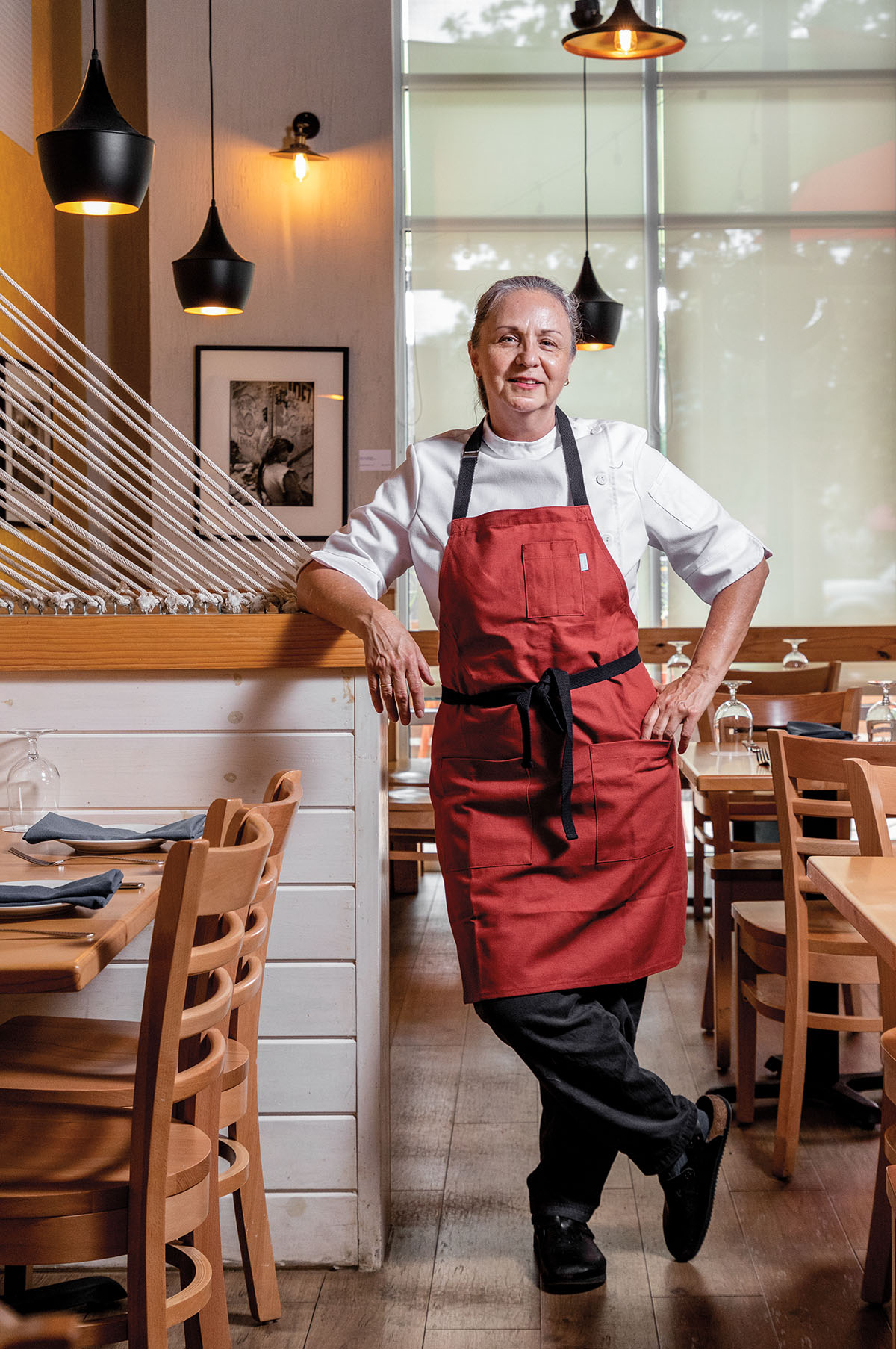 A woman in a red apron leans against a wooden ledge in a restaurant