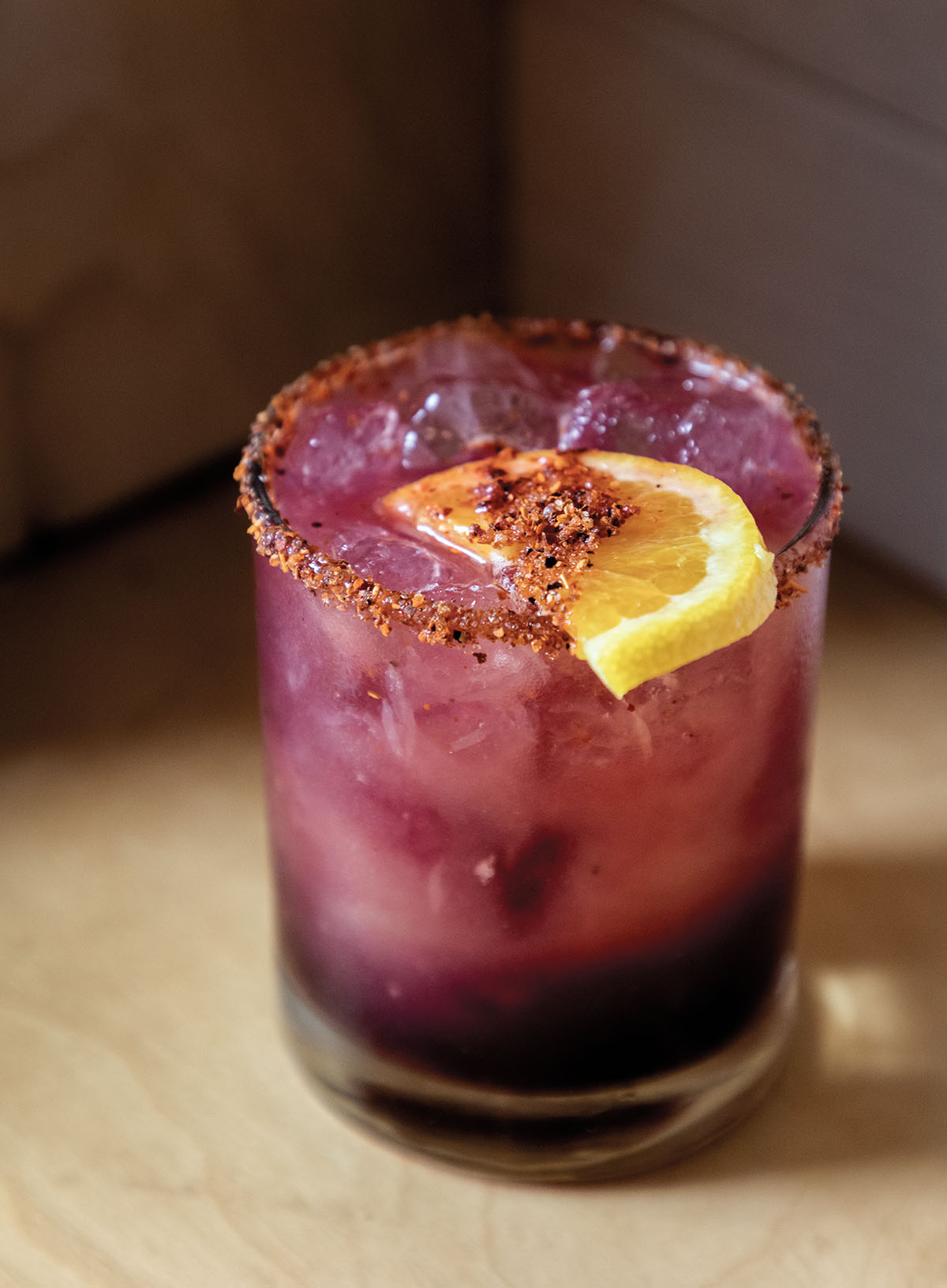 A purple cocktail with pieces of ice and a lemon wedge in a glass
