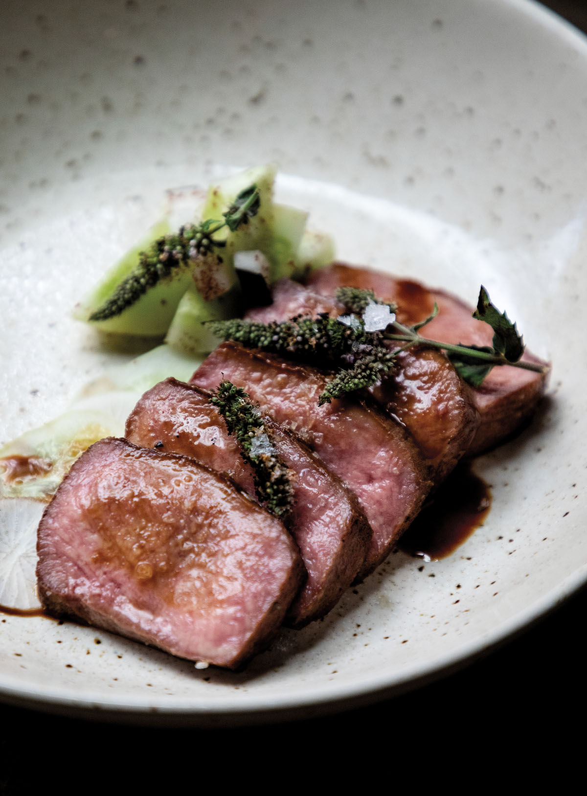 A plate with slices of red beef with a sauce and green garnish on a plate