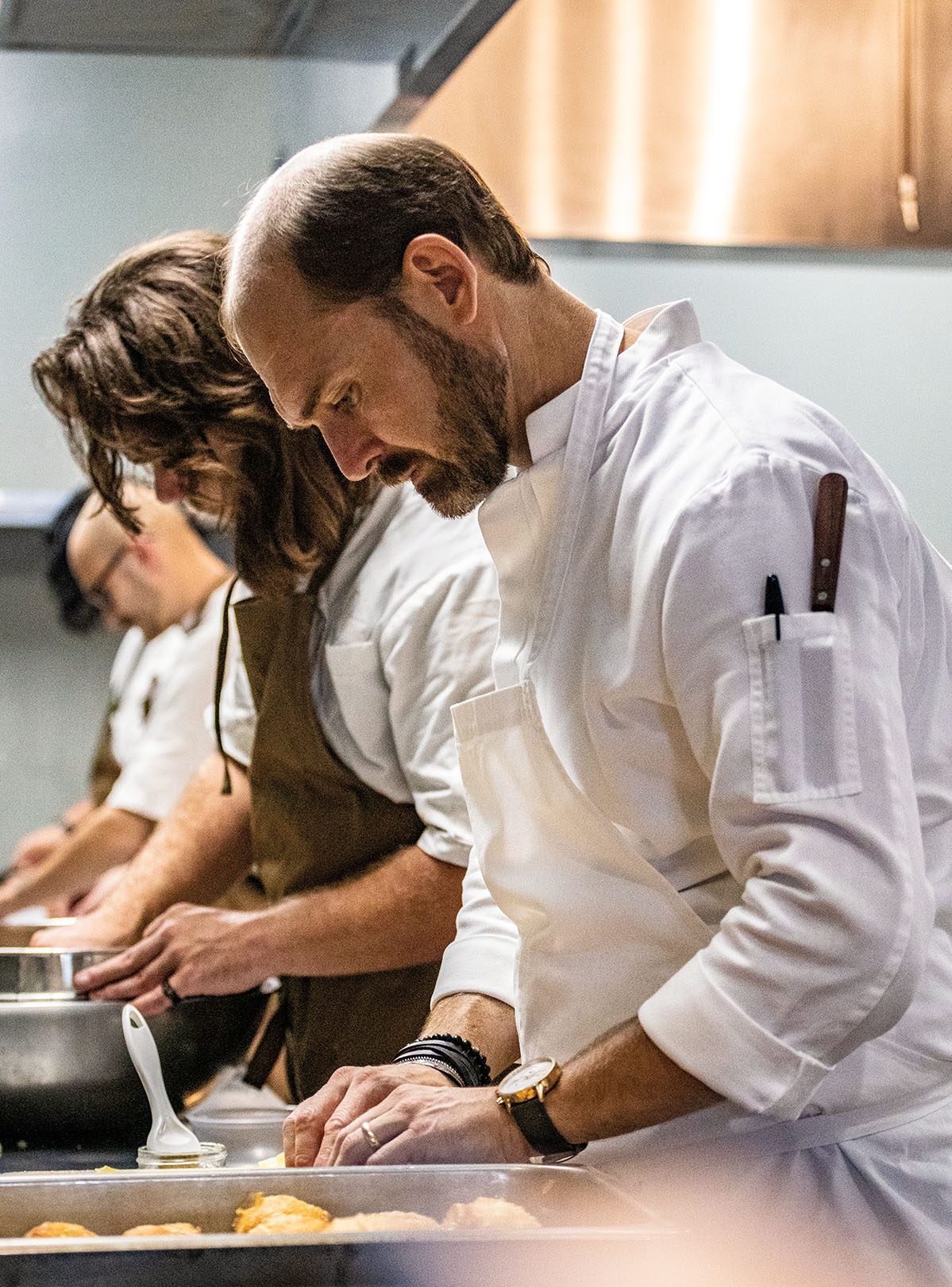 Three chefs work side by side in a kitchen in white chefs jackets carefully plating food in a well-decorated kitchen