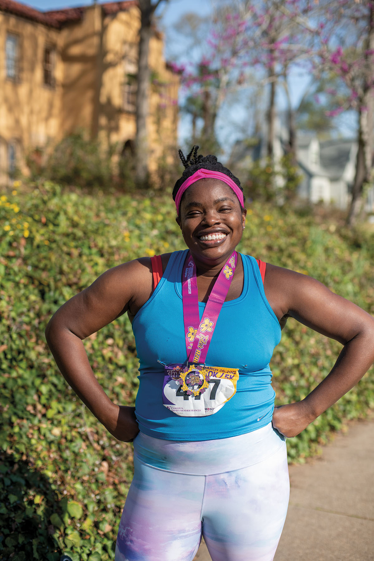 A woman wearing a blue tank top and pink sweatband stands with hands on hips and a race bib and medal on