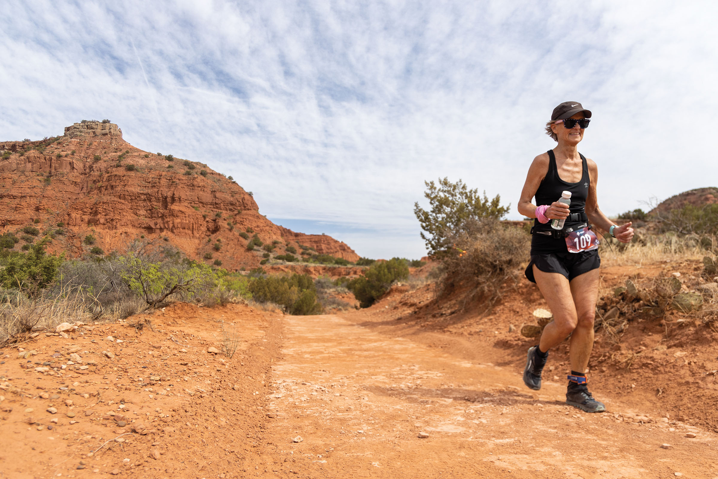 A woman in a black tank top and hat runs along a rugged dirt trail with canyons in the background