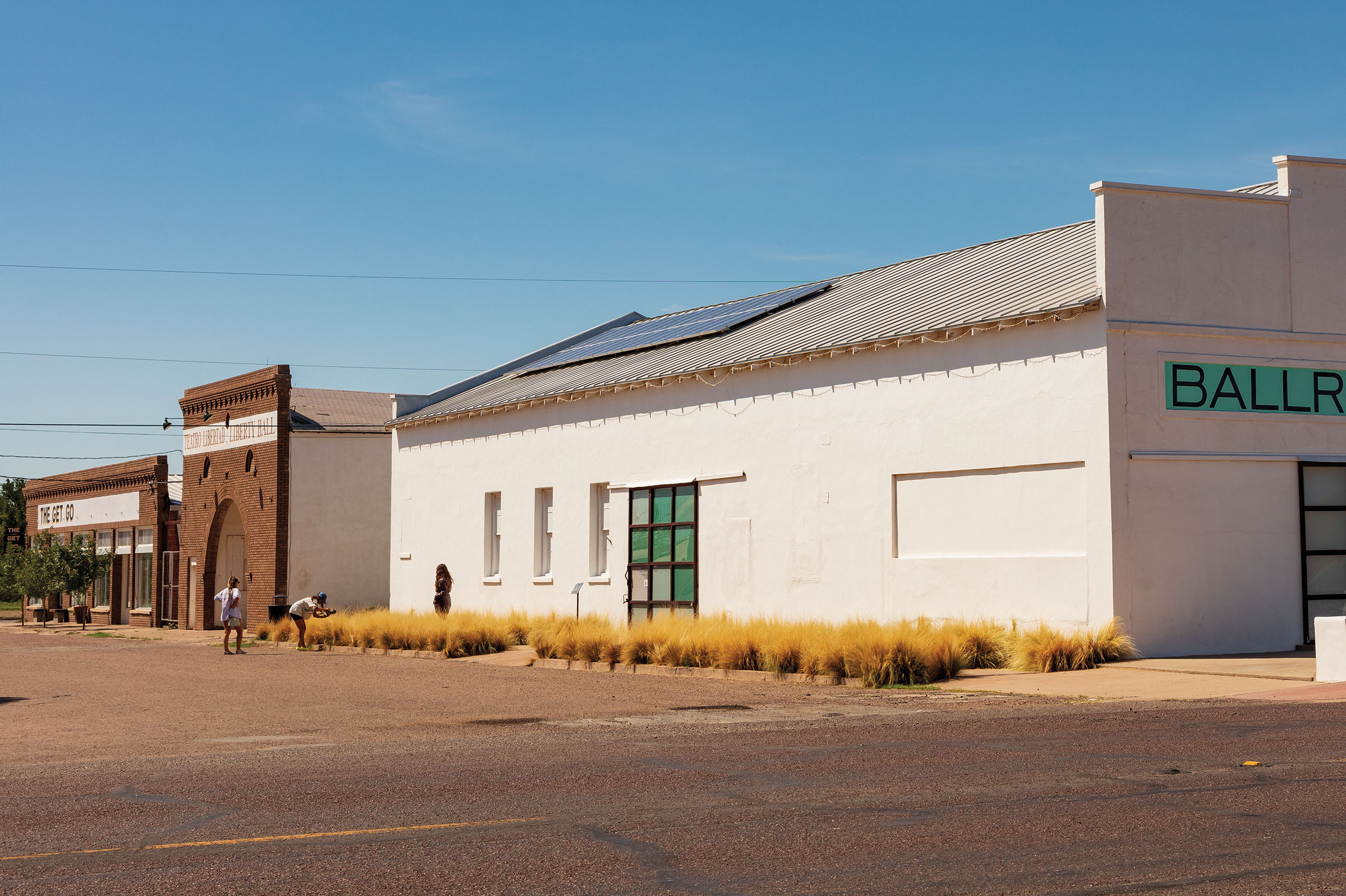 Two people pose for and take pictures in front of a large white building with solar panels under a cloudless blue sky