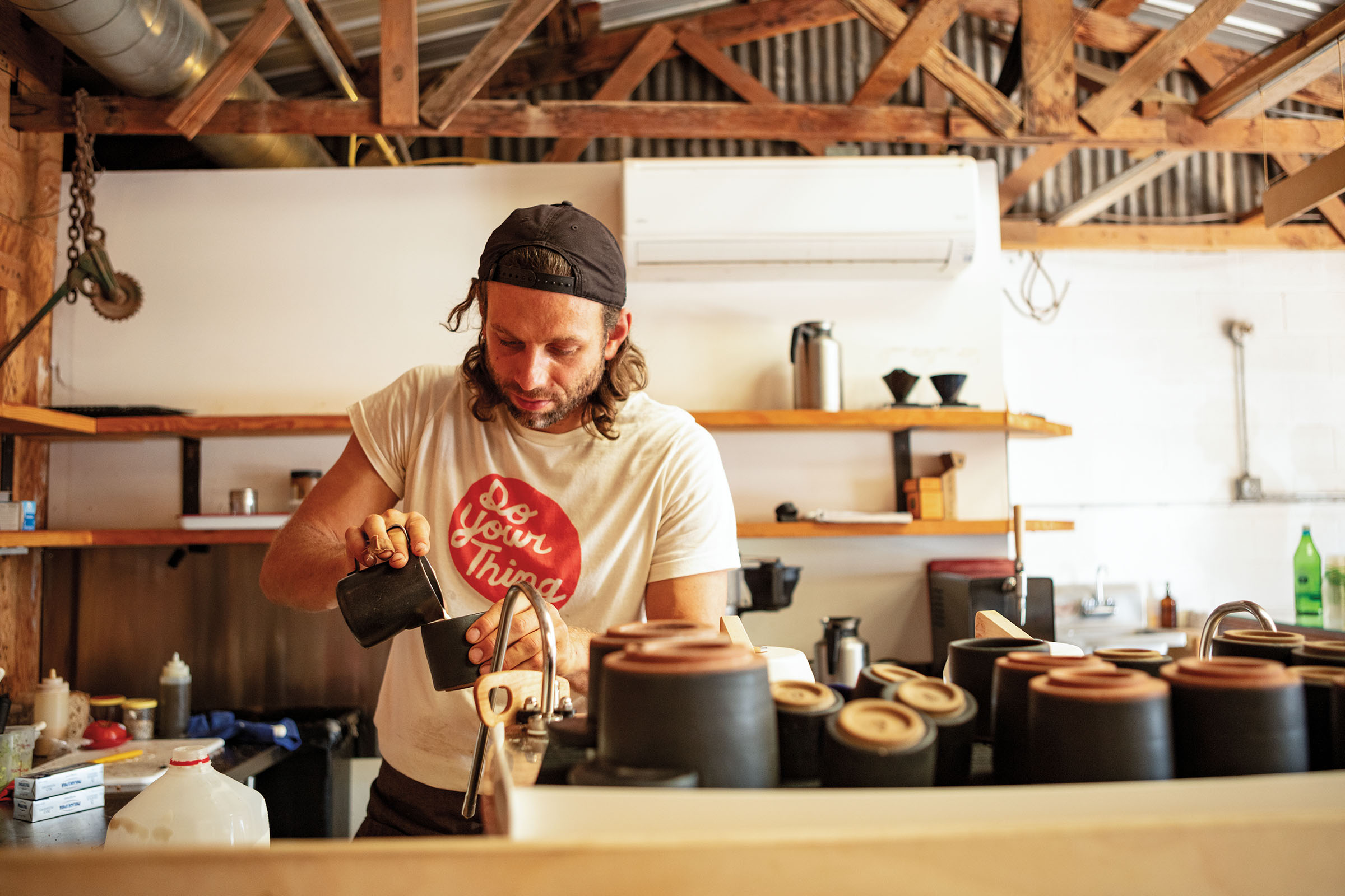 A man in a backwards baseball cap carefully pours milk into a coffee cup