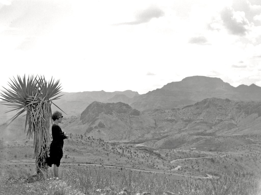 A black-and-white photograph of a woman leaning against a spiny cactus plant, with numerous mountains in the background