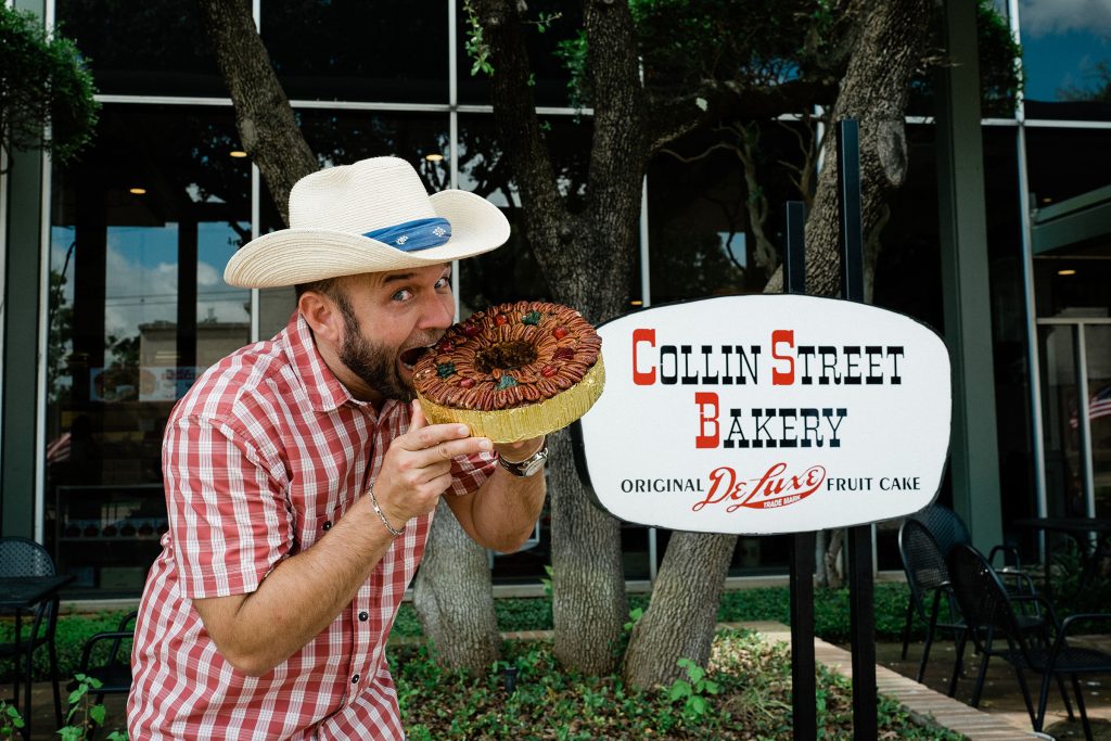A man in a white cowboy hat takes a large bite out of a fruitcake in front of a sign reading Collin Street Bakery