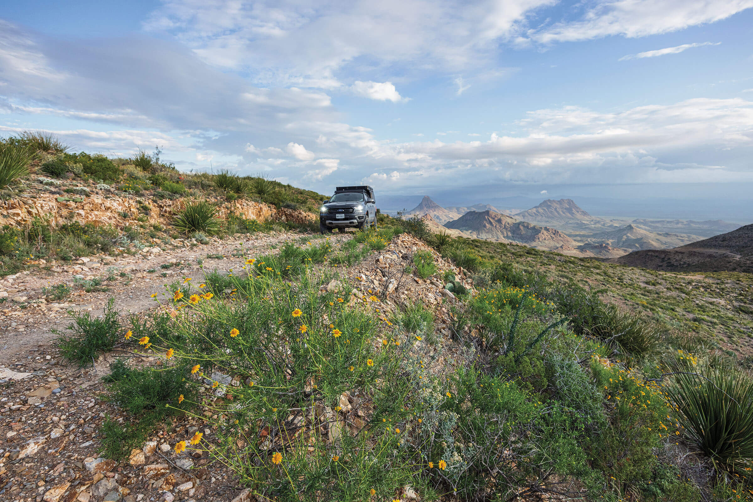 A car drives up a narrow track gravel road with a picturesque mountain scene behind