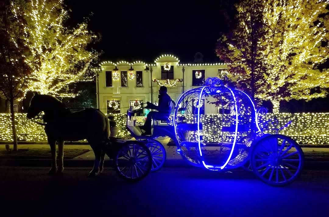 A round Cinderella-like horse-drawn carriage aglow with blue lights goes by a large two story historic home decorated with white Christmas lights and wreaths in Highland Park.