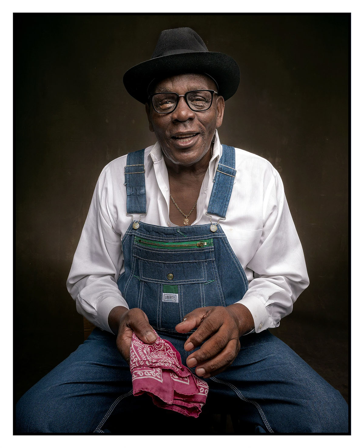 A portrait of a man wearing denim overalls holding a red cloth and wearing a black top hat