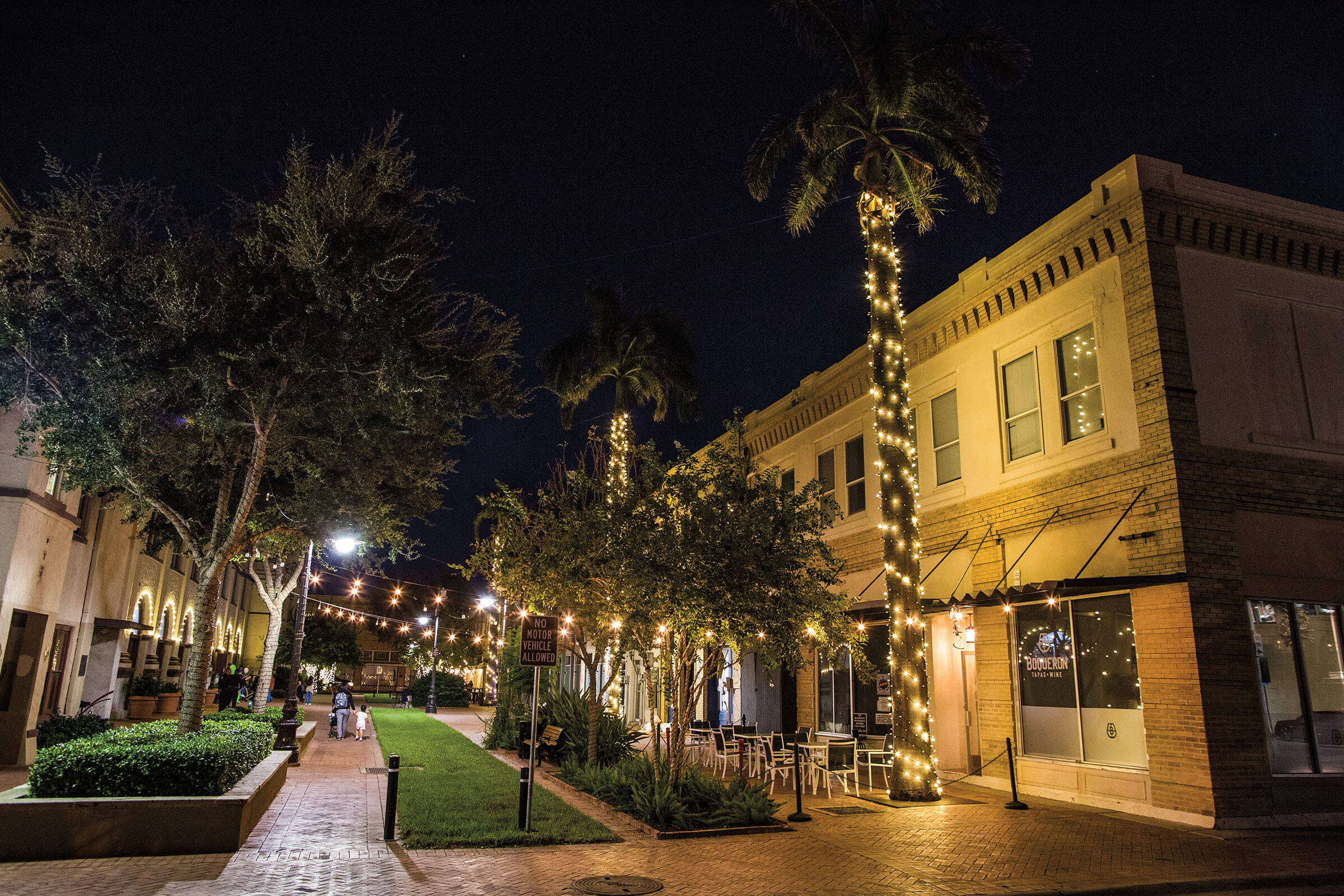 A downtown plaza with brick buildings adorned with glowing lights and people walking by