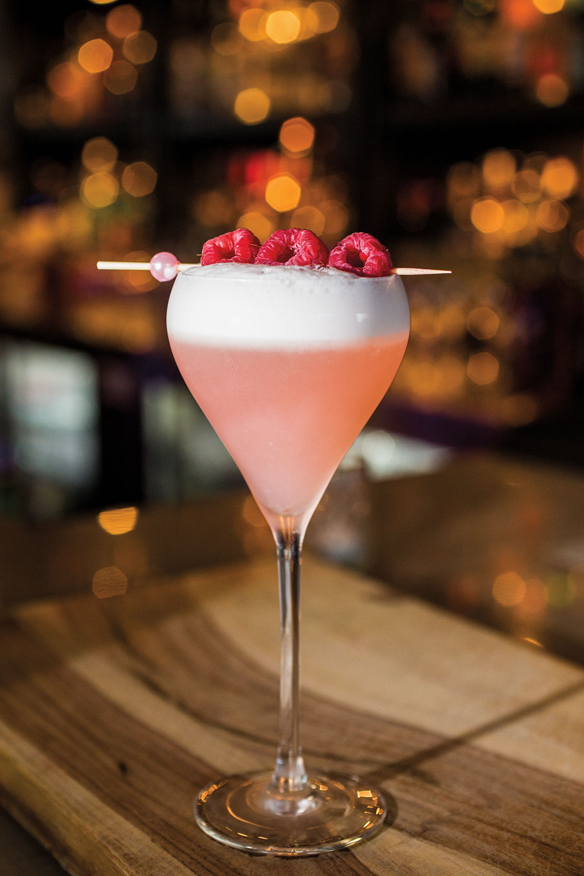 A rose-colored drink topped with fresh raspberries in a teardrop-shaped glass