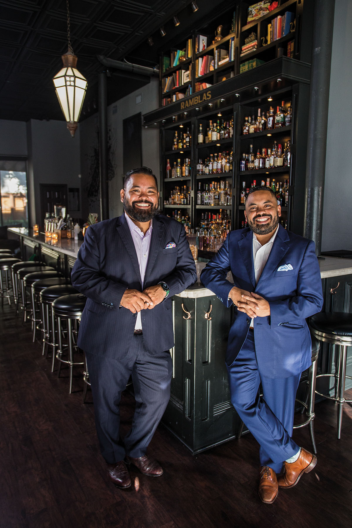 Two men in suits stand in front of a well-stocked bar with rich wood paneling