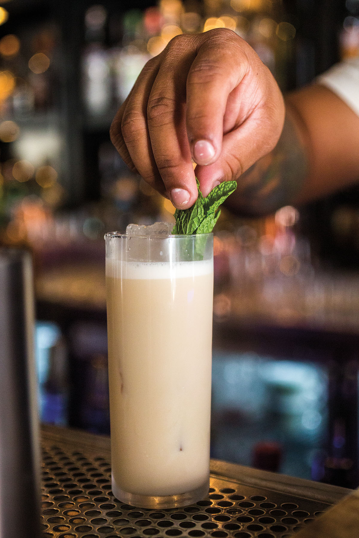 A man places a mint sprig in a tall class full of lightly opaque liquid