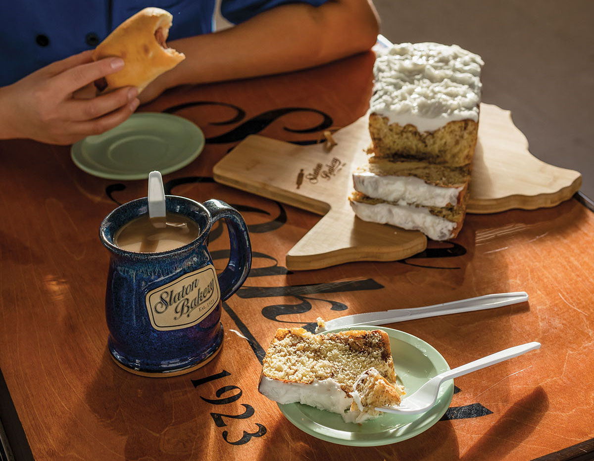 A spiced cake with white icing on a wooden table next to a cup of coffee