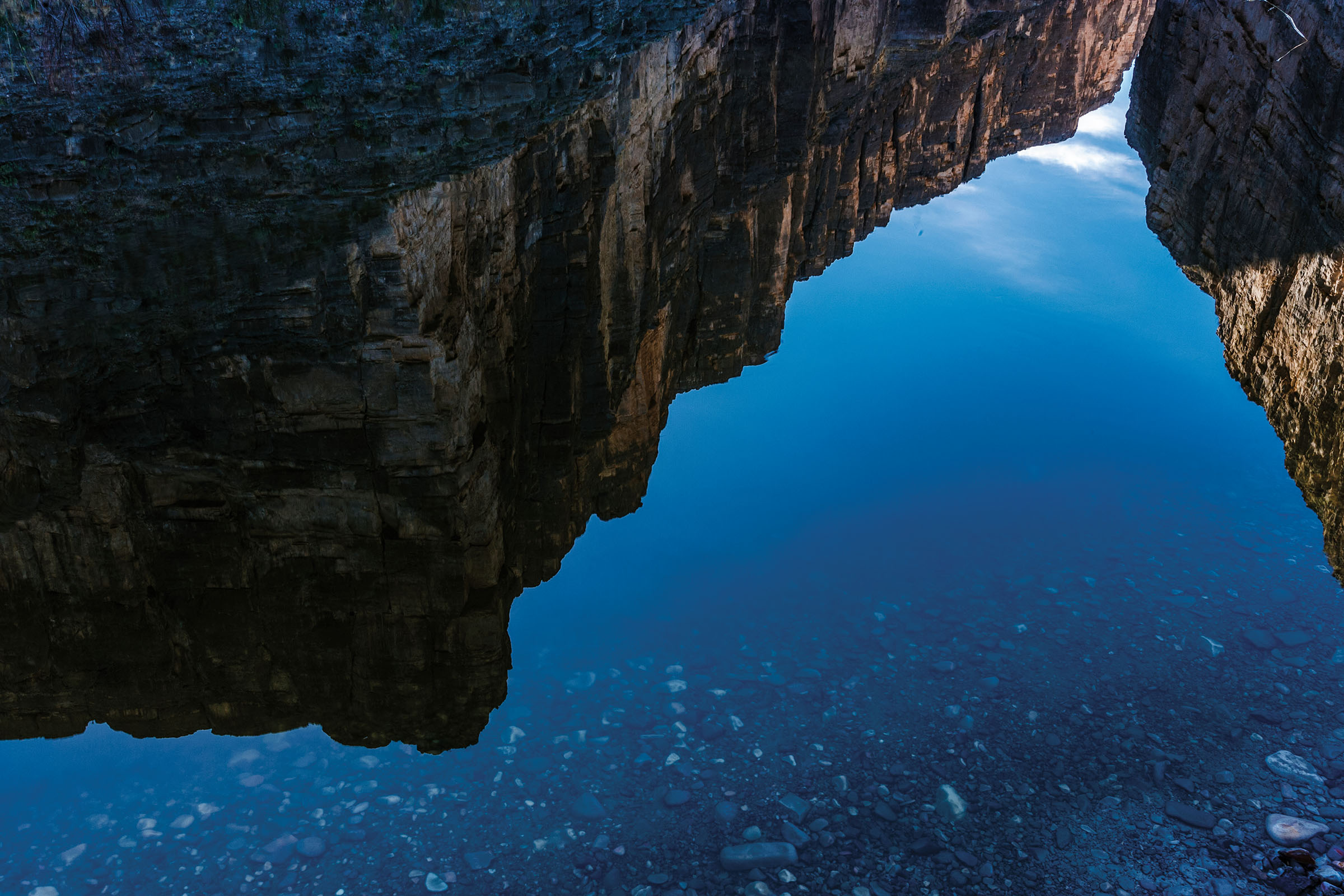 The reflection of a tall rocky canyon wall over blue water