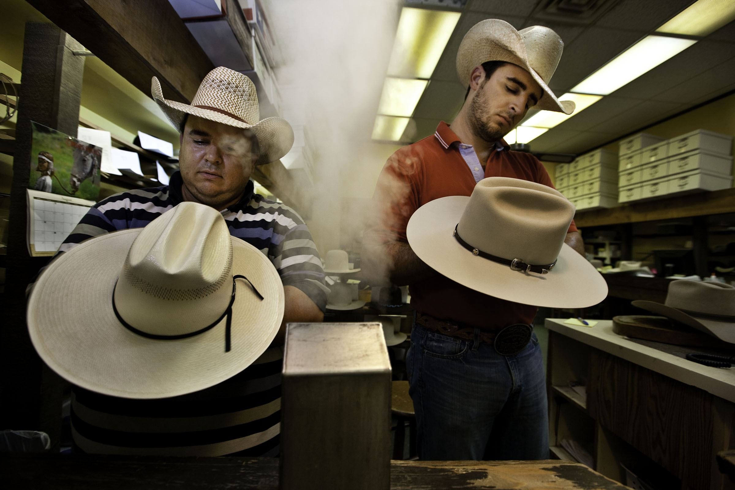 Two men wearing cowboy hats use steam to shape an additional two cowboy hats