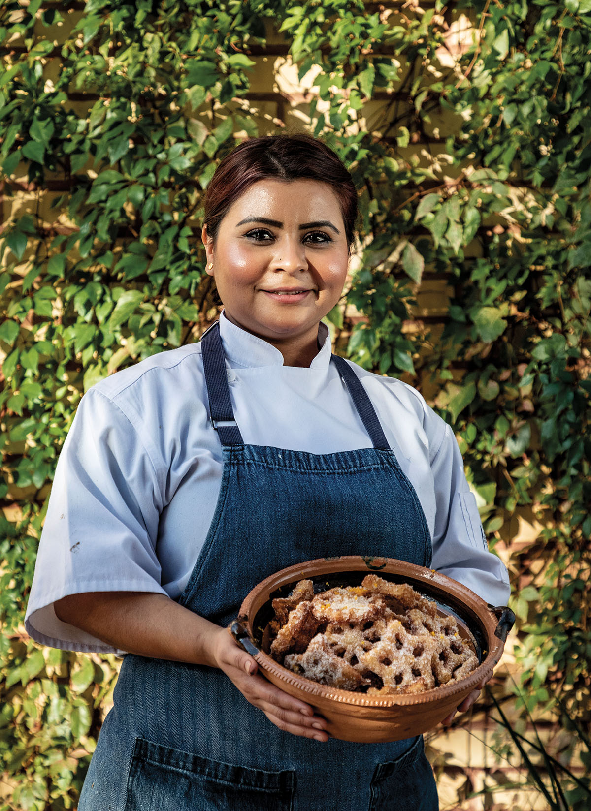 A woman in a blue collared shirt wearing a dark blue apron stands with a basket full of cinnamon sugar treats outdoors