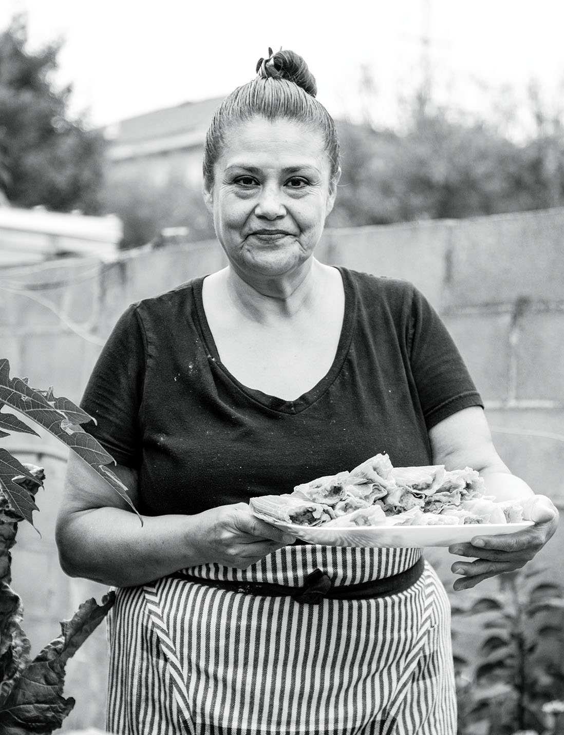 A woman wearing a pinstripe apron with her hair in a bun stands holding a large platter of tamales