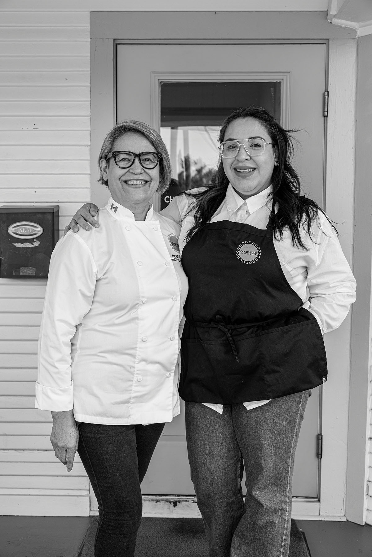 Two women, one in a chef's jacket and the other in a black apron, stand in front of a door