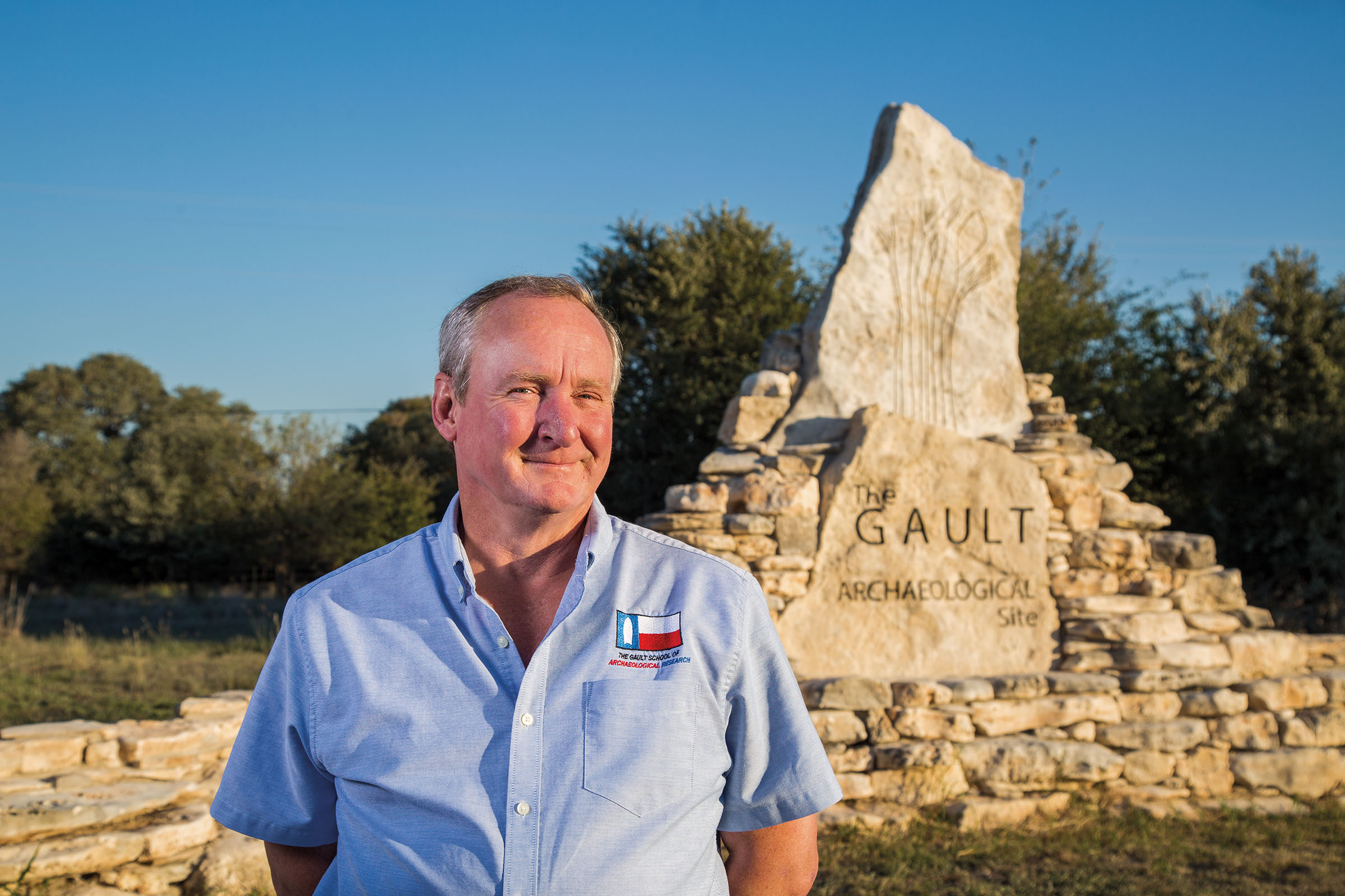 A man in a blue collared shirt stands in front of a rock formation with the word "Gault" etched into the side
