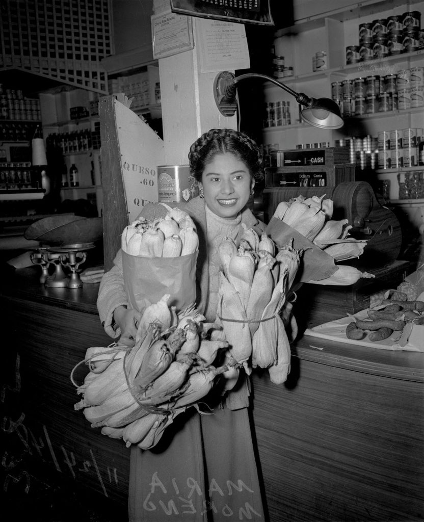 A black and white photograph of a woman holding armfuls of tamales