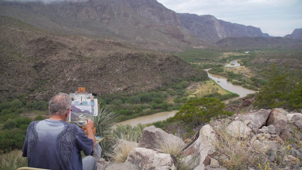 A man paints a picture of a park with a winding river and tall mountains