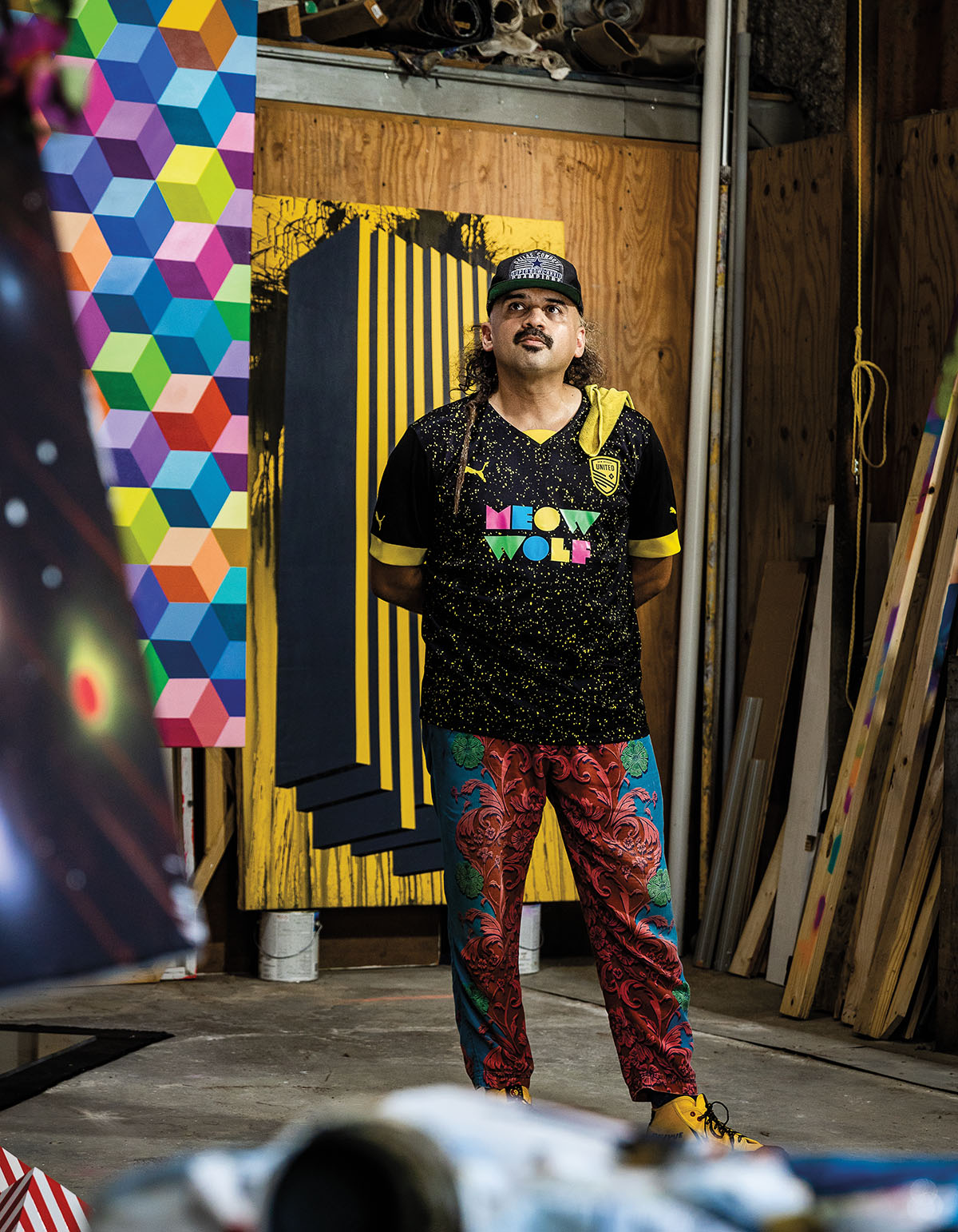 A man in a meow wolf t-shirt stands in a messy, colorful warehouse