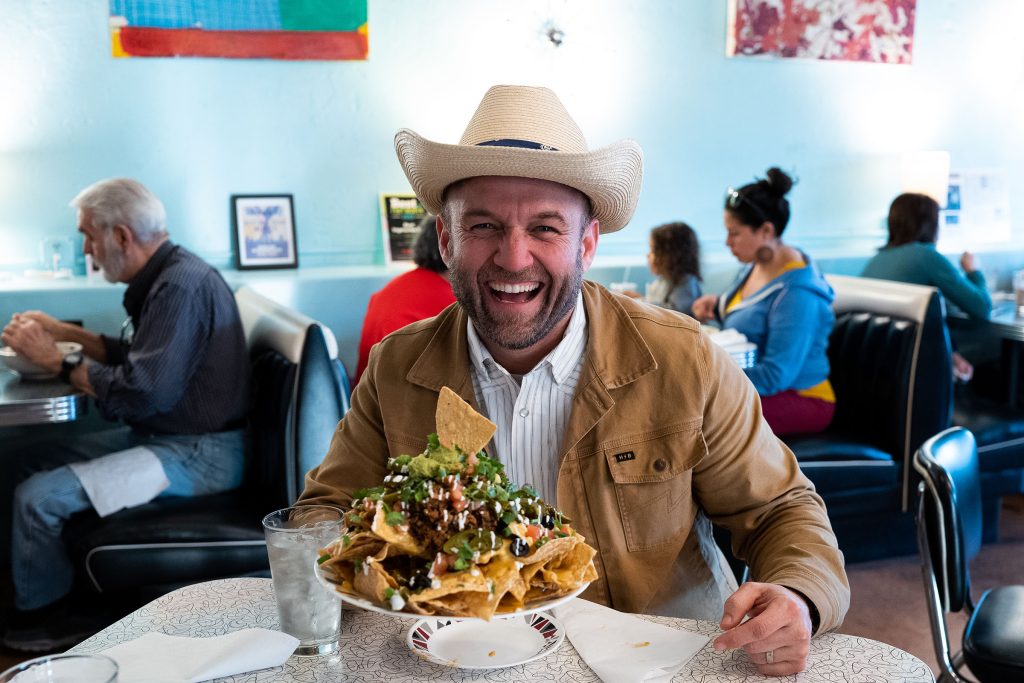 A man in a cowboy hat sits and laughs at a table behind a large platter of nachos