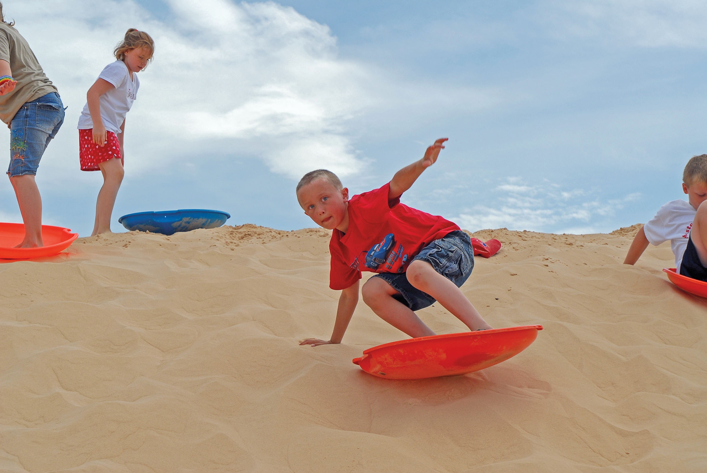 A young person in a red shirt surfs down a sand hill on a plastic saucer