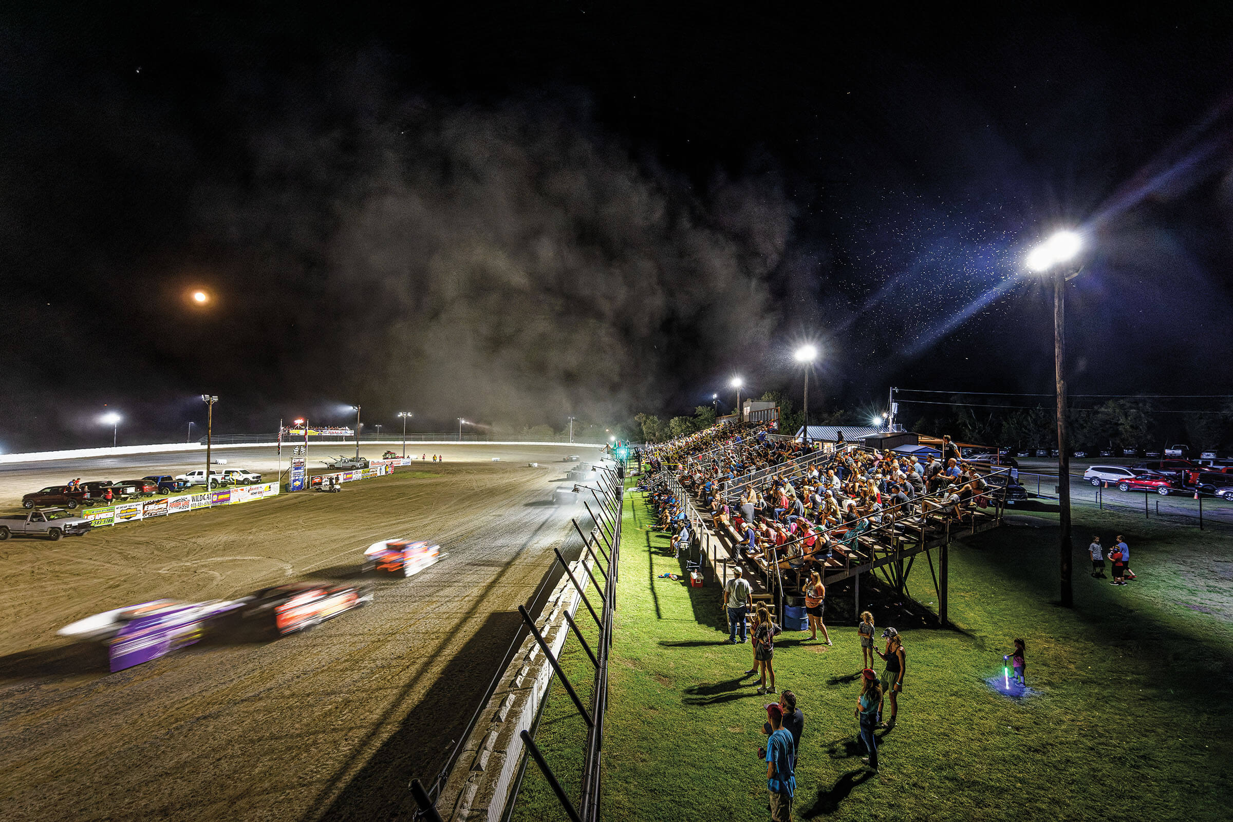 A crowd of spectators sit in stands as a group of racecars whiz by on a dirt track to their left