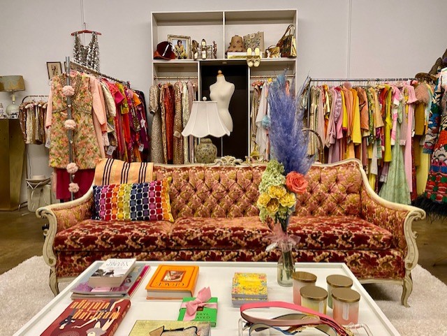 Interior of Evergirl Vintage shows a sofa with a coffee table strewn with old magazines and trinkets. Behind the sofa are clothing racks with vintage items. 