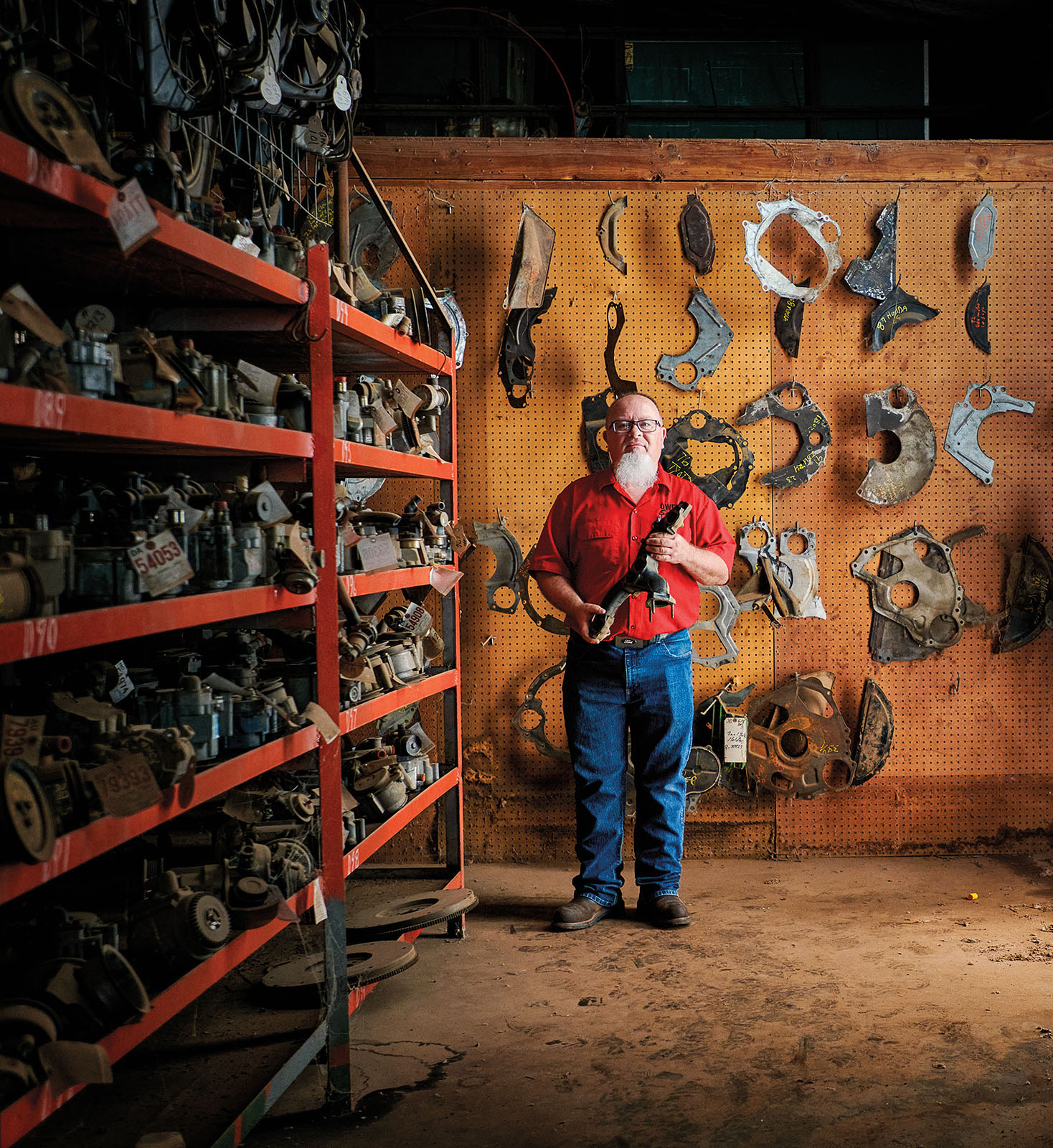 A man with a white beard wearing a red shirt and blue jeans stands in a well-organized warehouse of parts holding a piece of metal