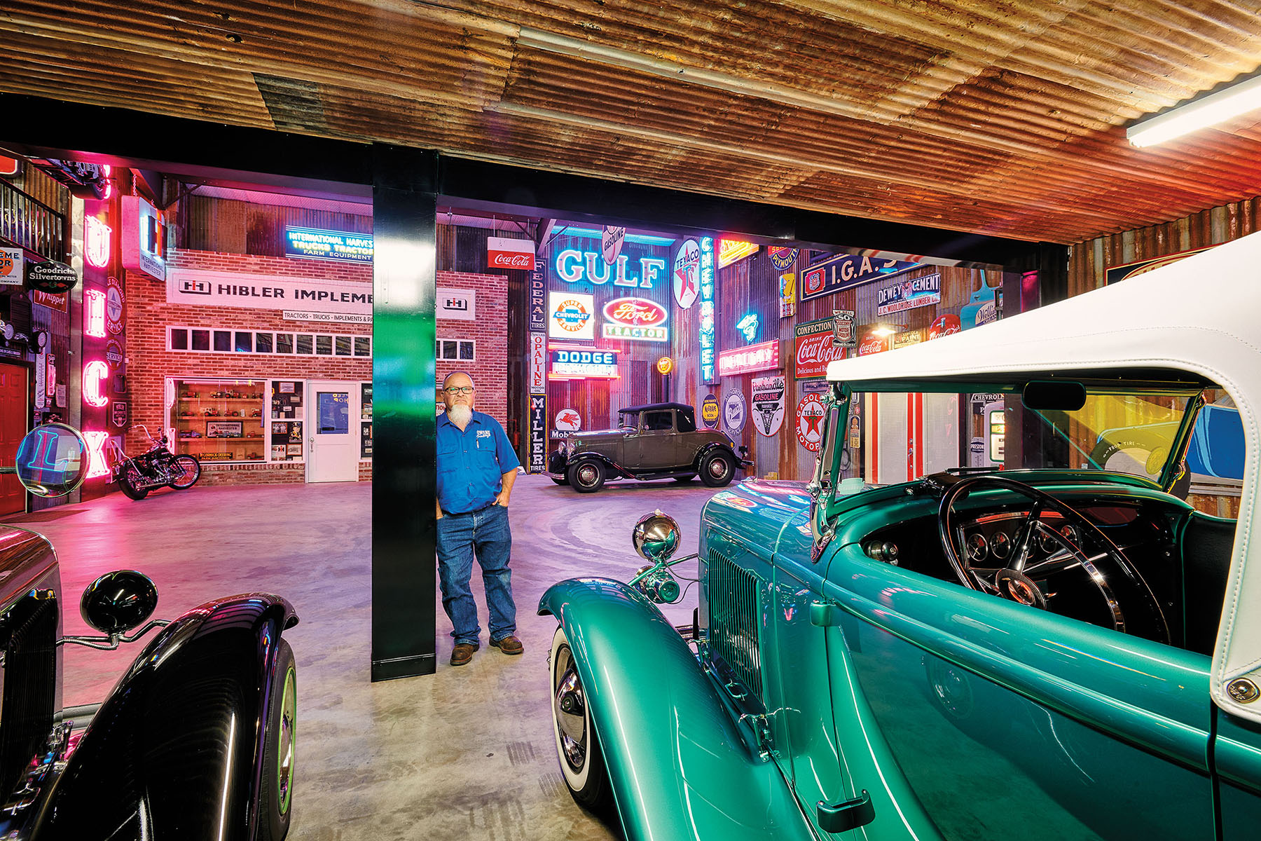 A man in a blue shirt and jeans leans against a metal post in a showroom with numerous bright neon signs