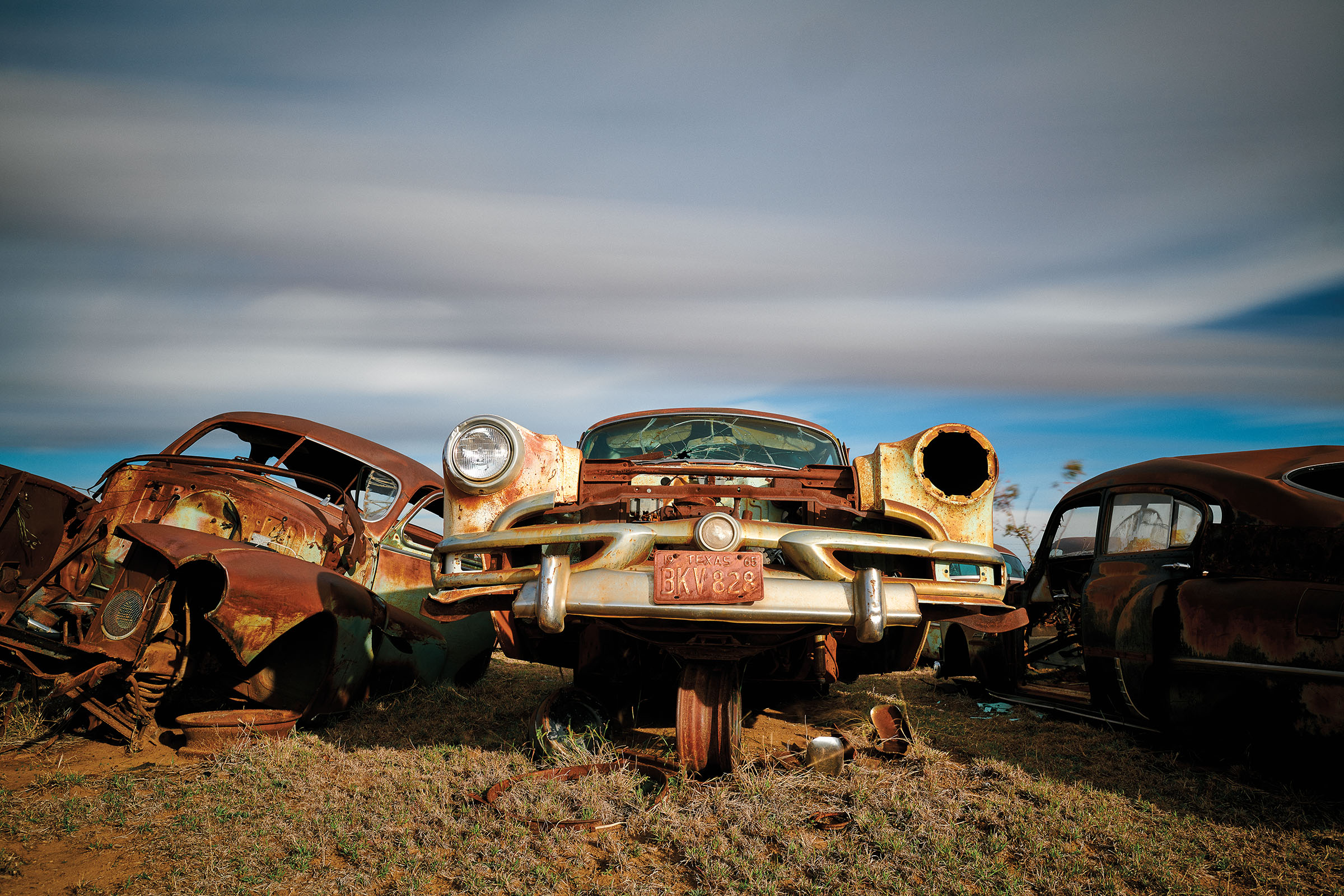 The hood of a rusted and old white car on a dirt plath under blue sky