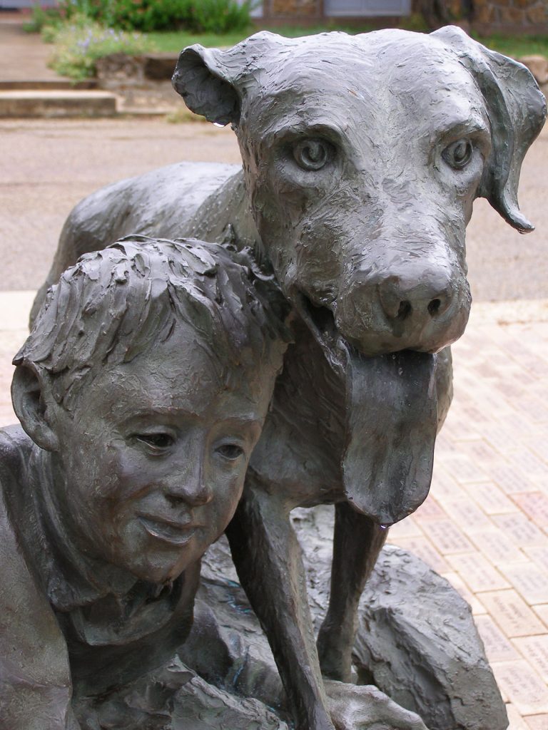 A close-up photo of the bronze statue, with young Travis Coates nestled against his dog, Old Yeller, whose tongue is hanging out of his mouth like he's panting.