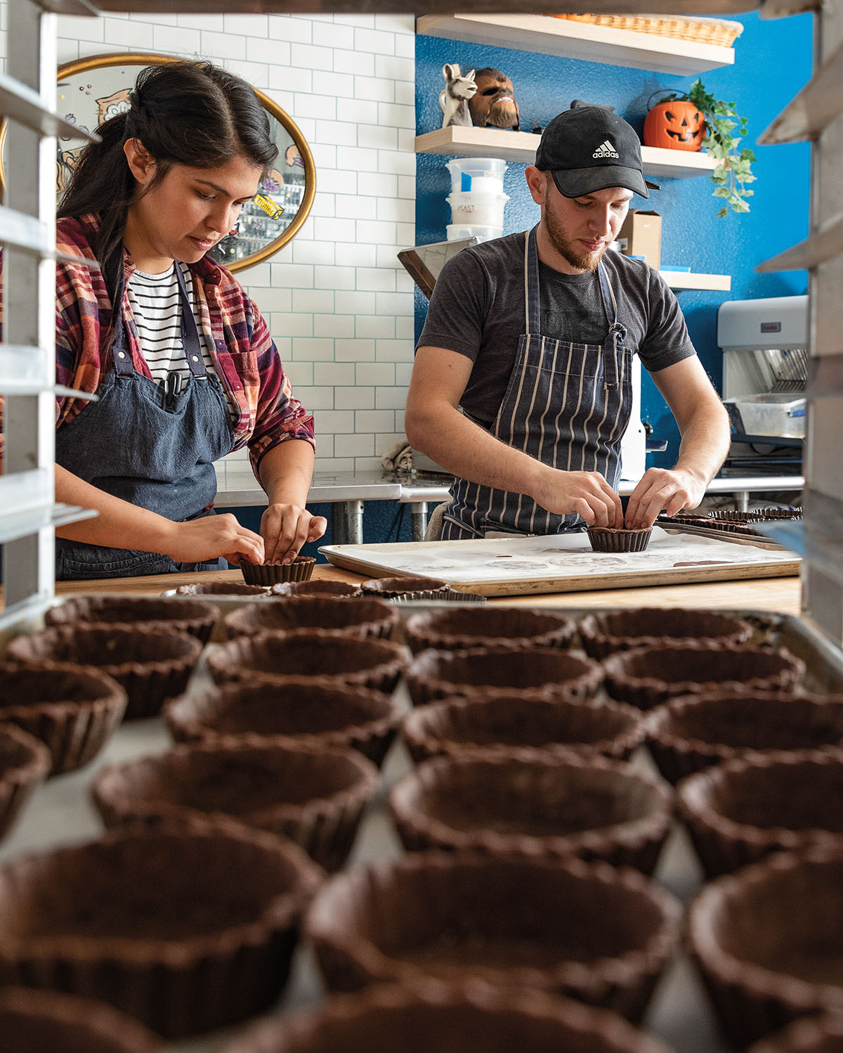 Two people stand in an industrial kitchen behind a tray full of chocolate cupcake liners