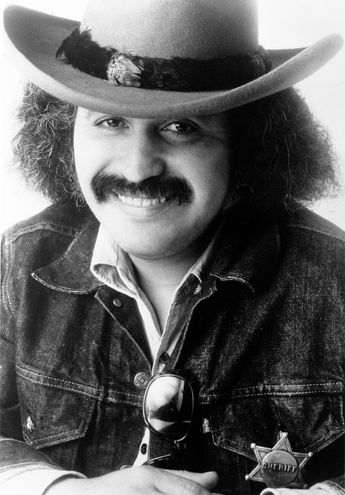 A man in a cowboy hat with long hair smiles in a black and white moustache