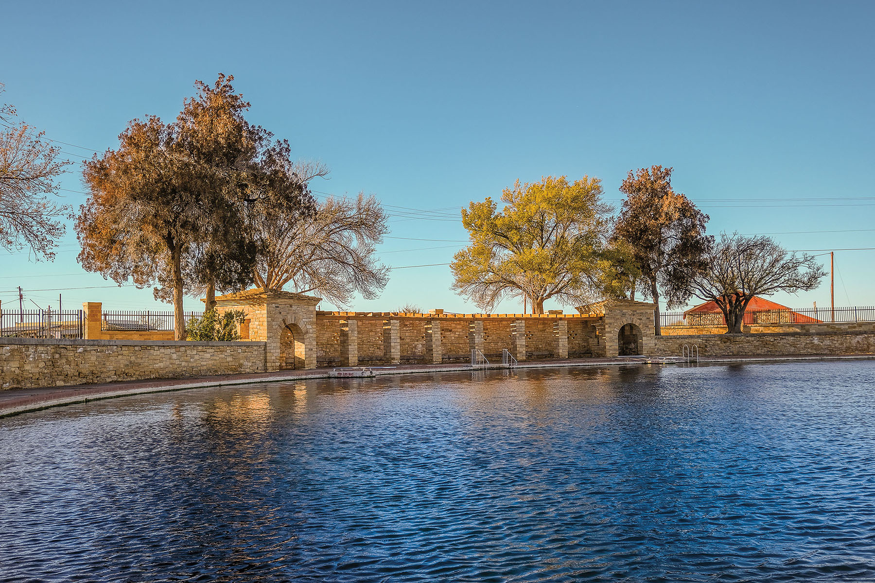 Golden light reflects across clear blue water onto a stone building next to tall green trees