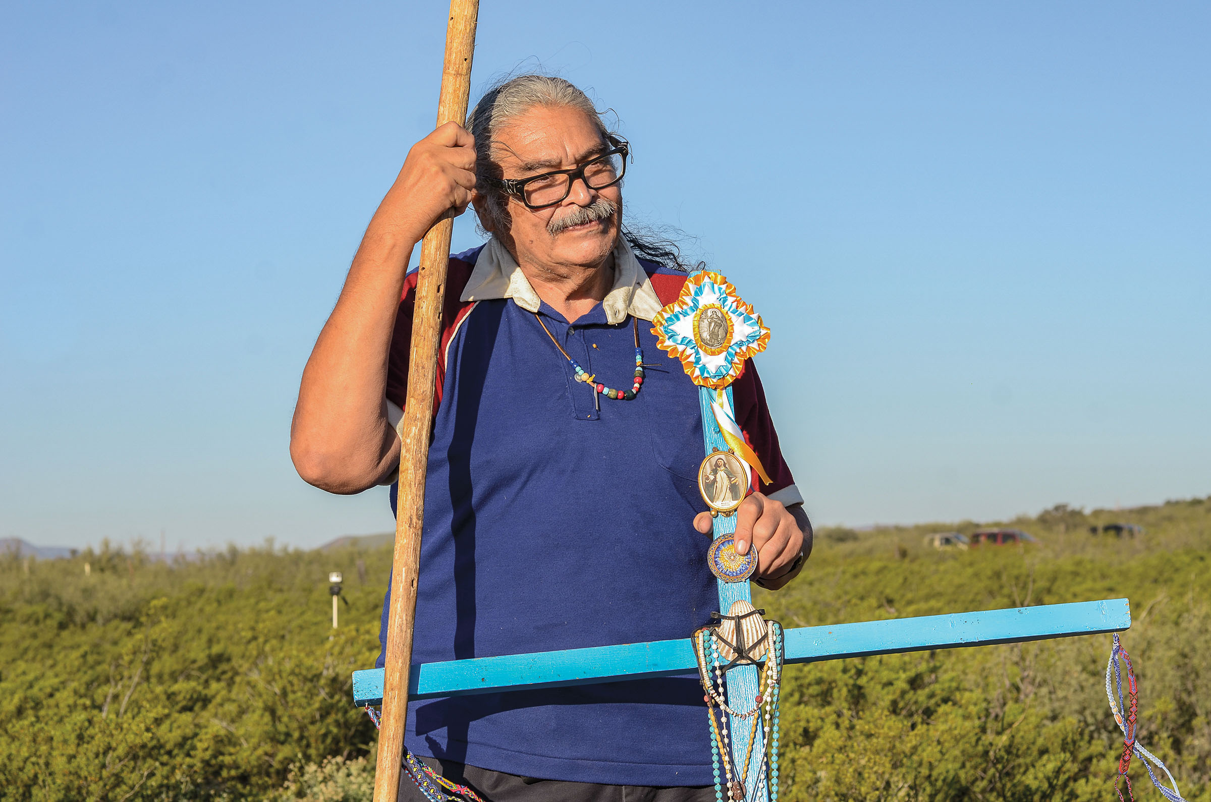 A man stands in a field with a turquoise cross decorated with beaded jewelery and intricate stitched designs