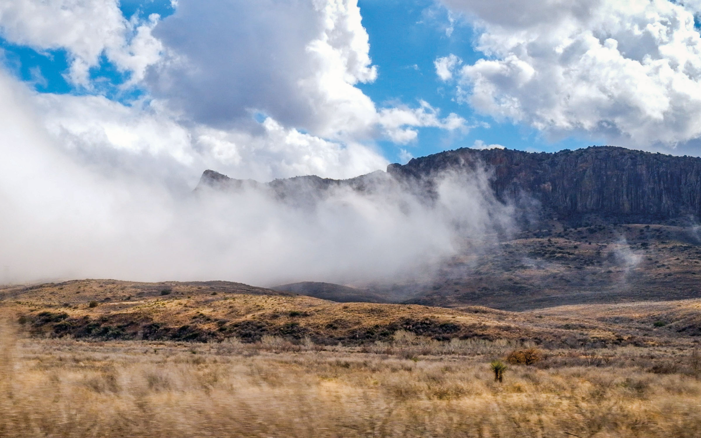 White fog rises up from a pool in rolling golden hills beneath tall blueish mountains