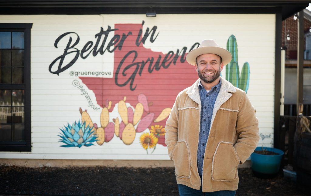 A man in a cowboy hat stands in front of a mural reading "Better in Gruene"