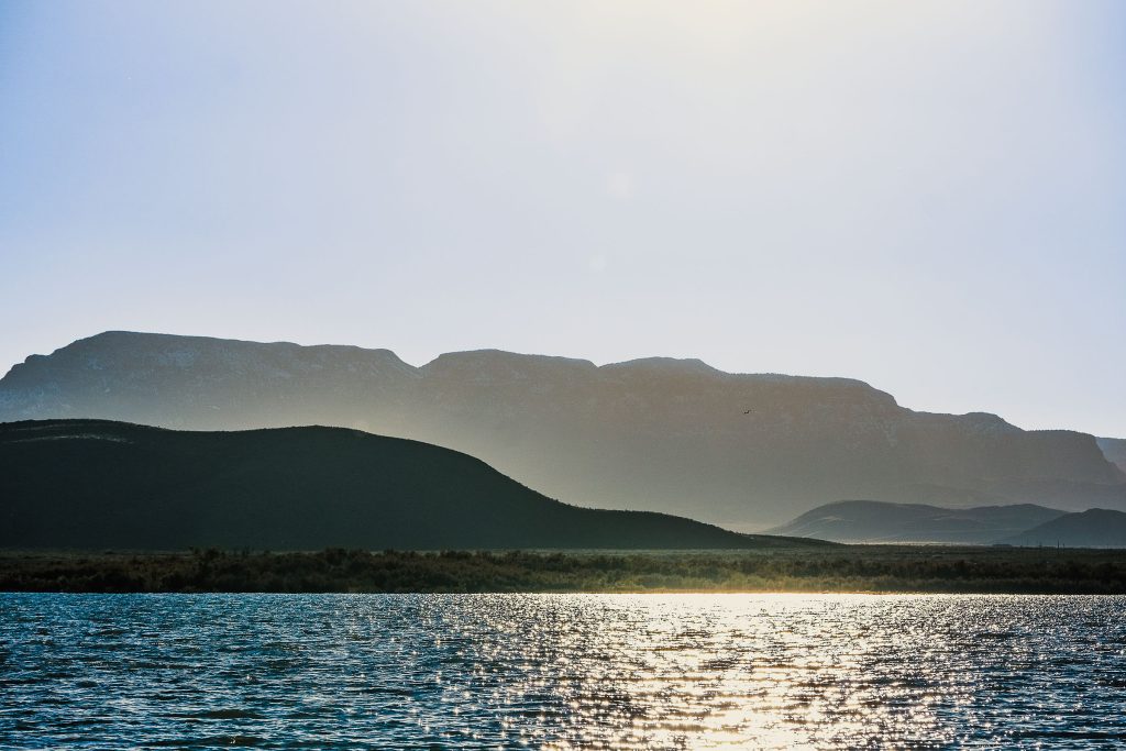 Tall mountains accentuate a large clear body of water under blue sky and sun